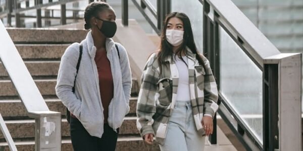 Full body young multiethnic female students in protective masks walking down stairs in campus and having conversation
